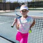 Smiling girl holding a tennis racket and ball on a sunny day at an outdoor tennis court.
