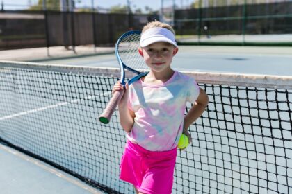 Smiling girl holding a tennis racket and ball on a sunny day at an outdoor tennis court.